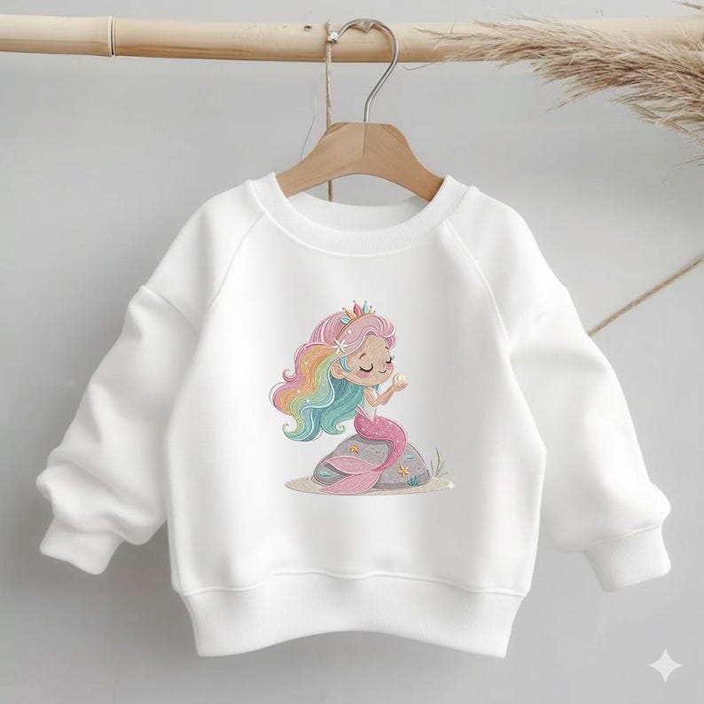 May include: White sweatshirt featuring a colorful mermaid illustration. The mermaid has rainbow hair, a pink tail, and a crown. The graphic is on a white, long-sleeved sweatshirt, perfect for casual wear.