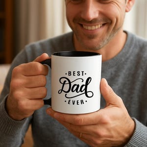 Puede incluir: Taza de cerámica blanca con interior y asa negros. La taza presenta el texto "BEST. Dad EVER." en una elegante escritura negra. La taza es sostenida por una persona que lleva un suéter gris.