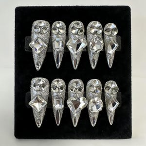 May include: A set of ten silver-glittered, stiletto-shaped false nails. Each nail is adorned with a large, clear, faceted gemstone in various shapes, including squares, diamonds, and floral designs. The nails are displayed on a black velvet surface.
