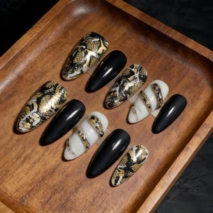Puede incluir: Colección de uñas artificiales en varios diseños, incluyendo negro, blanco y dorado. Algunas uñas presentan un patrón de piel de serpiente, mientras que otras son negras sólidas o tienen un diseño de espiral dorado. Las uñas están dispuestas en una bandeja de madera.