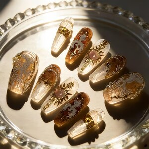 Puede incluir: Una variedad de uñas artificiales largas y ornamentadas. Las uñas presentan diseños en oro, marrón y blanco, incluyendo soles, fénix y patrones florales. Algunas uñas tienen bandas doradas y detalles de perlas. Están dispuestas sobre una bandeja plateada.