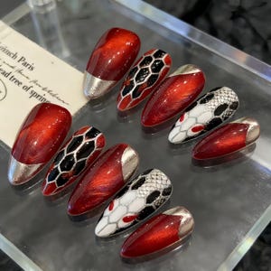 Könnte beinhalten: Ein Set aus zehn künstlichen Nägeln mit glänzend rotem Finish. Einige Nägel haben silberne Spitzen, während andere schwarz-weiße Schlangenhautmuster aufweisen. Die Nägel sind auf einer transparenten Oberfläche angeordnet.