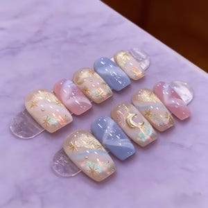 May include: A set of artificial nails with a celestial theme. The nails feature a variety of designs, including gold glitter, stars, and a crescent moon. Colors include light blue, pink, and gold. The nails are arranged on clear plastic holders.