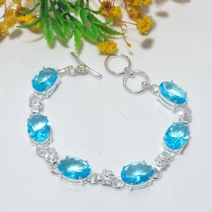 May include: A silver link bracelet featuring six oval-cut, bright blue gemstones set in prong settings. The bracelet includes small silver floral accents between each stone and secures with a silver toggle clasp consisting of a bar and a ring.