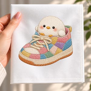 May include: A white fabric square featuring an embroidered design of a small, round, cream-coloured chick with pink cheeks sitting inside a colourful patchwork trainer. The trainer has pastel-coloured panels, beige laces, and a tan rubber sole.