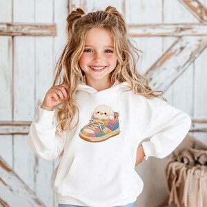 May include: A white long-sleeve hooded sweatshirt featuring a central graphic of a small, round, light yellow chick sitting inside a colourful, patchwork-style trainer with tan laces and a textured brown sole, designed with a front kangaroo pocket.