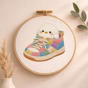 May include: A circular wooden embroidery hoop containing a white fabric panel with a stitched design of a small white bird with pink cheeks sitting inside a colourful patchwork sneaker. The sneaker features pastel pink, blue, yellow, and green panels.