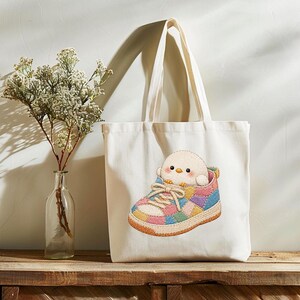 May include: A cream-coloured canvas tote bag featuring an embroidered graphic of a small, round, white bird sitting inside a colourful patchwork trainer. The trainer has laces and a multi-coloured pattern of pink, blue, yellow, and orange squares.