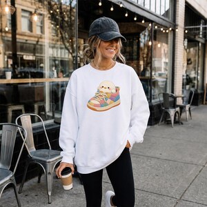 May include: A white crewneck sweatshirt featuring a graphic of a small yellow chick sitting inside a colourful patchwork sneaker. The sneaker has a textured, multi-coloured pattern with light laces and a tan sole, centred on the chest of the garment.