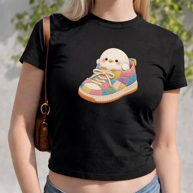 May include: A black short-sleeved t-shirt featuring a graphic of a small, round, cream-coloured bird sitting inside a colourful patchwork trainer. The trainer has a textured appearance with multi-coloured panels in shades of pink, blue, and yellow.