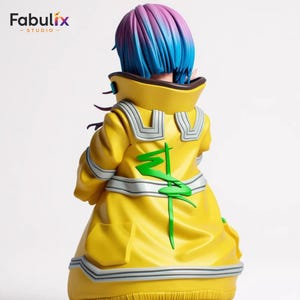 May include: A figurine of a character with blue and purple hair, wearing a bright yellow coat with green and silver accents. The coat has a high collar and a stylized green design on the back. The Fabulix Studio logo is in the top left corner.