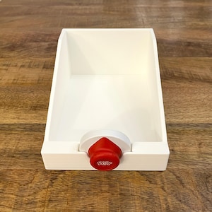 May include: A white open-top storage bin with a slanted back and sides. The front features a circular red knob centered on the edge, which has the word PLAY embossed in white letters on its surface. The bin has a smooth, matte finish throughout.