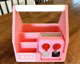 May include: A pink plastic caddy with a white handle, featuring two open storage compartments, a nameplate labeled Sarah, and an integrated pink electronic device with two red knobs, a speaker grille, and a digital display showing 11:00.