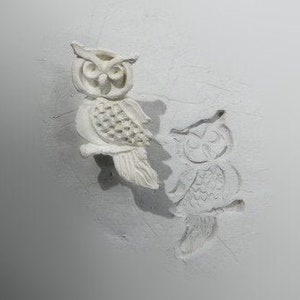 May include: A white ceramic owl figurine with a detailed design. The owl is perched on a white surface and has a shadow cast behind it. There is a second, smaller owl design imprinted in the surface behind the figurine.