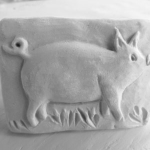 May include: A rectangular, off-white relief sculpture of a pig. The pig is depicted in profile, with its curly tail and pointed ears visible. The pig stands on a bed of stylized grass. The sculpture is a monochromatic piece.