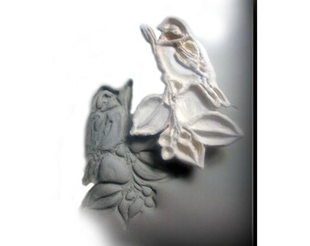 May include: A white ceramic wall sculpture of two birds perched on a branch with leaves. The sculpture is in a minimalist style and has a textured surface.