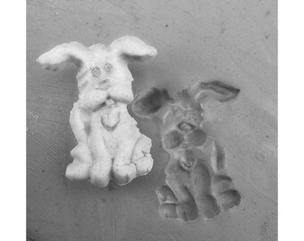 Może przedstawiać: Dwie małe, białe ceramiczne figurki królików. Jeden królik siedzi prosto z uszami uniesionymi do góry, podczas gdy drugi królik siedzi z głową pochyloną w dół. Oba króliki mają szczegółowe rysy twarzy.