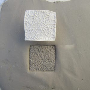 May include: A white ceramic tile with a raised floral design is placed on top of a gray clay tile with the same design impressed into it.