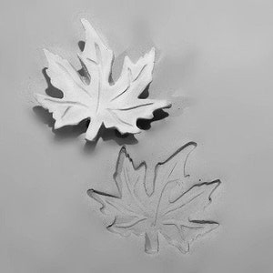 Pode incluir: Duas decorações de folhas de bordo brancas, uma em relevo e outra embutida, contra um fundo cinza. As folhas têm veias detalhadas e uma aparência tridimensional, sugerindo um tema inspirado na natureza.