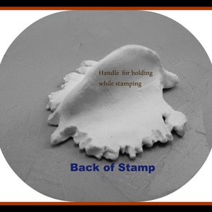 May include: A close-up of the back of a white clay stamp with a handle for holding while stamping. The text "Back of Stamp" is printed below the stamp.