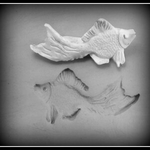 May include: A white ceramic mould of a fish, with a raised impression of the fish below it.