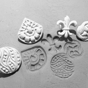 May include: A collection of white plaster decorative elements, including a fleur-de-lis, a round floral design, and various other embossed shapes. The objects are arranged on a textured, light-colored surface.