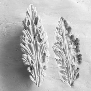 May include: Two white plaster leaf-shaped moldings with intricate details. The moldings are on a white textured background.