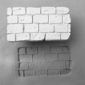 May include: A white brick wall texture stamp, with a textured impression of the stamp on a gray surface below.
