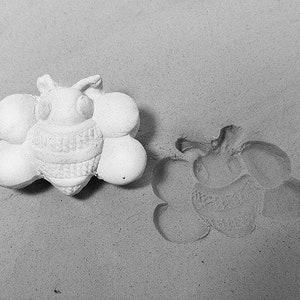 May include: A white ceramic mold of a bee with a textured body. The mold is surrounded by smaller, round molds. The bee mold is slightly raised from the surface.