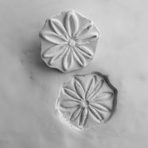 May include: Two white clay flower stamps with a raised design. The stamps are pressed into a white clay surface.
