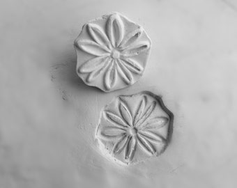 May include: Two white clay flower stamps with a raised design. The stamps are pressed into a white clay surface.