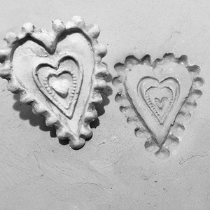 May include: Two heart-shaped, off-white decorative pieces with textured surfaces. Each heart features an inner heart design and a scalloped edge. The pieces are set against a light gray background.