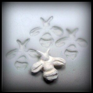 May include: A white ceramic mold with four bee shapes pressed into the surface. A single, raised bee is sitting on the mold.