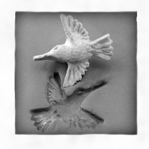 May include: A white ceramic hummingbird sculpture with its wings spread, perched on a gray surface. The sculpture is casting a shadow of itself on the surface.