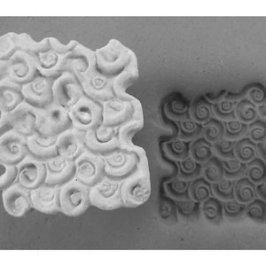 May include: A close-up of a white ceramic tile with a raised design of swirling patterns. The tile is next to a gray impression of the same design in a soft material.