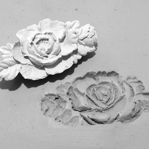 May include: Two white plaster roses, one with a more detailed design than the other, are shown in a close-up view.
