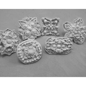 Pode incluir: Seis botões decorativos de cerâmica branca com designs florais intrincados. Os botões são de várias formas e tamanhos, incluindo redondos, quadrados e em forma de estrela.