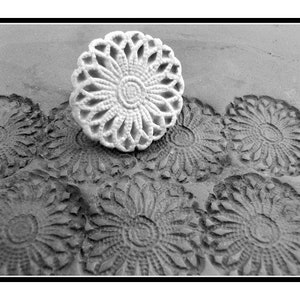Op de afbeelding: Een witte, decoratieve, ronde kantonderlegger met een bloemenmotief. De onderlegger ligt op een oppervlak met verschillende andere vergelijkbare onderleggers die in het oppervlak zijn gedrukt.