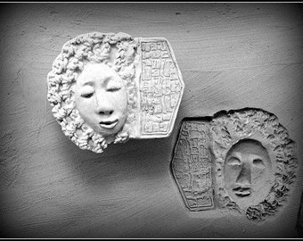 May include: Two black and white ceramic wall sculptures of faces with intricate details. The sculptures are mounted on a white wall.
