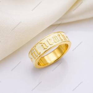 May include: A gold-toned band ring featuring an off-white enamel inlay. The ring is engraved with the text 'til death' in a black gothic-style font. The surface of the ring is smooth and polished, showcasing a metallic finish along the inner band.
