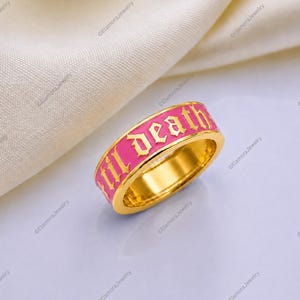 May include: A gold-toned band ring featuring a vibrant pink enamel inlay. The ring displays the text "Lil Death" in a raised, gothic-style gold font. The design is consistent around the outer circumference of the metal band, showcasing a bold contrast between the bright pink background and the metallic gold lettering.
