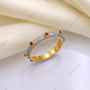 Könnte beinhalten: Ein goldener Bandring mit einer glatten grauen Emaille-Oberfläche auf der Außenseite. Der Ring ist mit fünf kleinen, runden, dunkelroten Edelsteinen verziert, die in regelmäßigen Abständen auf der Außenseite des grauen Emaille-Bandes eingefasst sind.