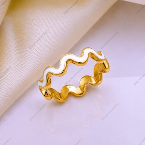 Puede incluir: Un anillo de tono dorado con un diseño continuo de ondas o zigzag. La superficie superior de la banda está incrustada con un acabado de esmalte blanco opaco y liso que sigue la forma ondulada del metal, creando un contraste distintivo.