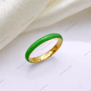 Puede incluir: Un anillo de oro con una banda de esmalte verde brillante. El anillo es de estilo sencillo, con un interior de oro pulido. El esmalte verde es de un color vibrante y sólido. El anillo se muestra sobre una superficie blanca con una tela blanca en el fondo.