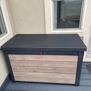 May include: A large outdoor storage box with a black top and a light brown, wood-look front panel. The box has a black frame and a red latch. The box is sitting on a dark gray deck.