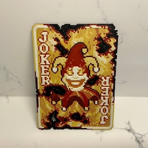 May include: A pixelated Joker playing card design. The card features a smiling jester with a red hat and collar, set against a yellow and brown background. The word "JOKER" is vertically displayed on both sides of the card.