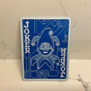 May include: A blue and white playing card featuring a pixelated jester design. The word "JOKER" is printed vertically on both sides of the card. The jester wears a hat and has a smiling face.