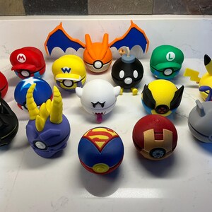 Op de afbeelding: Een verzameling van vijftien bolvormige figuren met verschillende karakterthema's, waaronder een rode Mario-pet, een gele Wario-pet, een groene Luigi-pet, een witte geest, een zwarte bom, een gele Pikachu en diverse superheld-geïnspireerde motieven.
