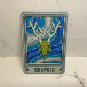 May include: A rectangular card featuring a pixelated illustration of a deer skull with large antlers. The skull is yellow-green, set against a blue and white background. The word "CRYPTID" is displayed at the bottom.