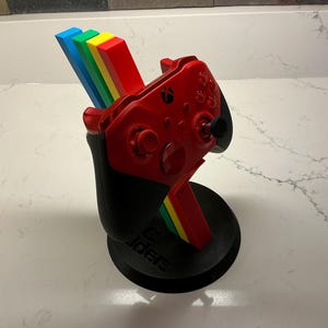 May include: A red Xbox controller is displayed in a black stand with a rainbow accent. The stand has a circular base and a curved support. The controller is red with black buttons and a joystick. The rainbow accent includes blue, green, yellow, and red.